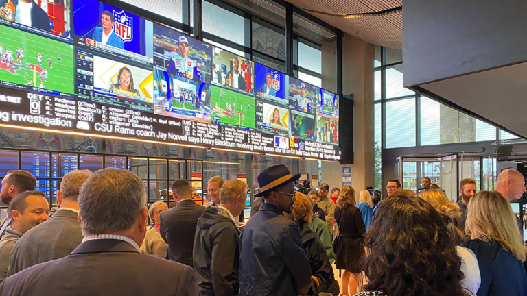 A crowded sports lounge featuring a large video wall displaying various sporting events. Numerous patrons are gathered in front of the screens, watching different games simultaneously.